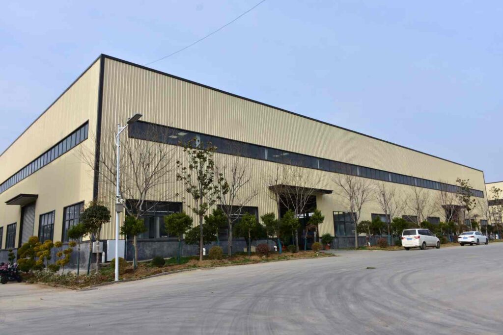 A panoramic photo of a modern industrial plant building, featuring a large main structure with corporate signage on the exterior walls, surrounded by well-maintained greenery under a clear sky.
